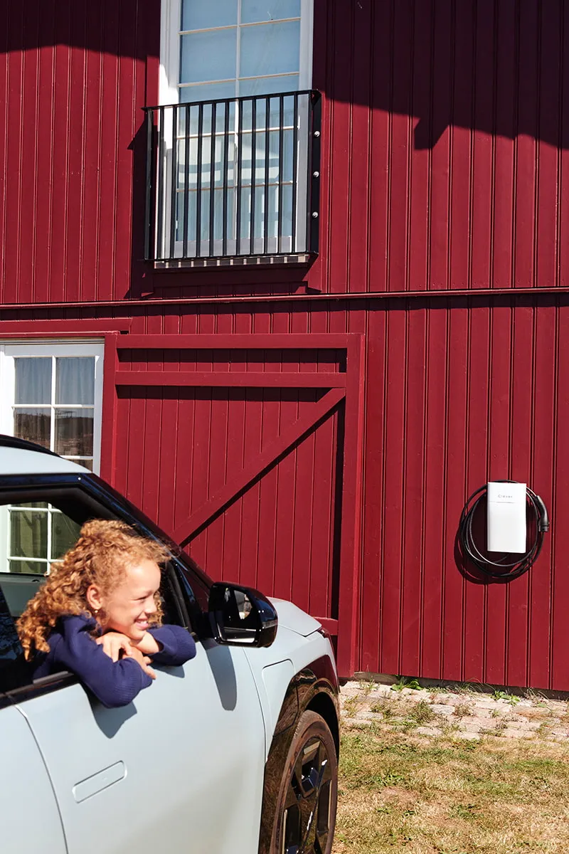Barn læner sig grinende ud af en elbil foran en rød bygning med Clever ladeboks monteret på væggen.