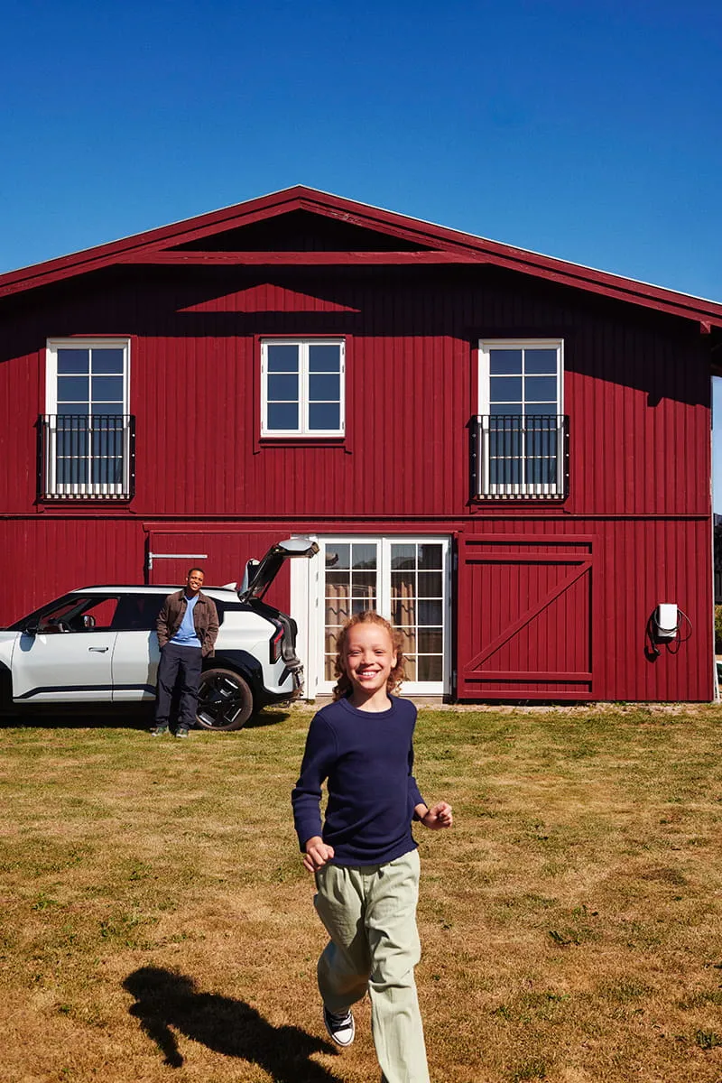 Barn løber på græsplænen foran en rød trævilla, mens en elbil holder ved Clever ladeboks på husmuren.